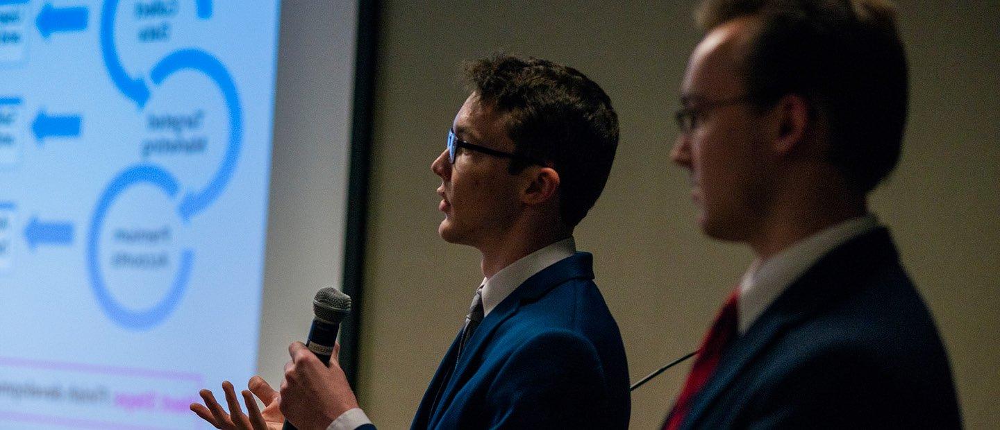 Two young men in suits, one holding a microphone, standing next to a projector screen.